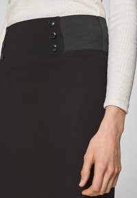 Černá sukně s vysokým pasem a šedým žebrovaným pruhem, zdobená pěti černými knoflíky na straně, vyrobená z hladkého, elastického materiálu.