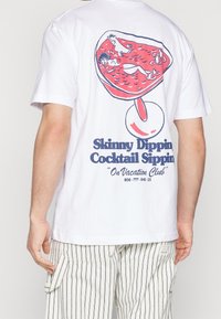 Biała koszulka z ilustracją czerwonego kieliszka koktajlowego, przedstawiająca dwie osoby pływające, z niebieskim napisem "Klub Wakacyjnego Picia Koktajli na Boso".