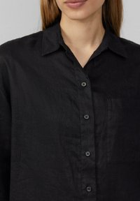 Camisa negra de lino con cuello, cierre frontal de botones y un solo bolsillo en el pecho. La tela tiene una apariencia texturada y un ajuste ligeramente holgado.