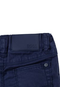 Pantalones de algodón elástico azul oscuro con un parche de cuero con logo, costuras visibles y un detalle de botón en el bolsillo. Textura suave y diseño ajustado.