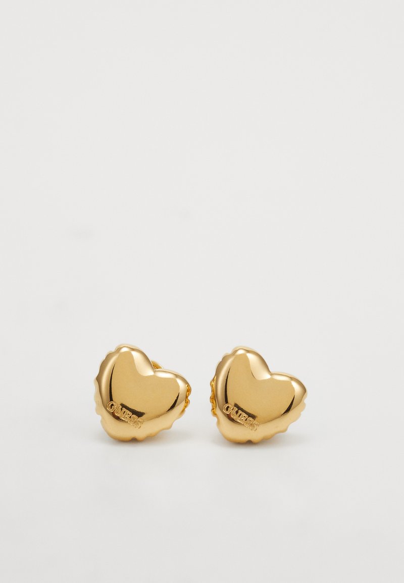 Gouden hartvormige oorbellen met een gladde, reflecterende oppervlakte en golvende randen, met aan de ene kant de merknaam "GUESS" gegraveerd.