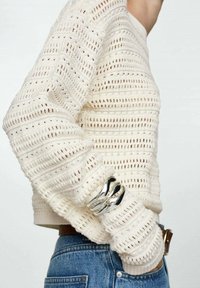 Maglione lavorato a maglia di colore crema con un motivo testurizzato, caratterizzato da un ampio scollo e maniche lunghe. Accentatura in argento sui polsi.