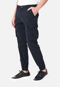 Pantaloni cargo blu navy con tasche laterali, polsini elasticizzati e una texture liscia; indossati con sneakers nere con suola bianca.