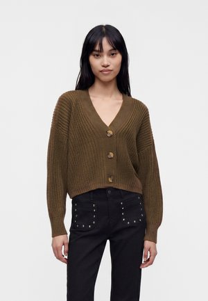 Jeune femme aux longs cheveux noirs porte un cardigan marron en tricot boutonné et un pantalon noir avec des poches avant cloutées.