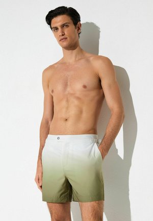 Bărbat fără tricou, sprijinit de un perete alb, purtând pantaloni scurți de înot cu un gradient verde și alb, cu mâinile în buzunare.