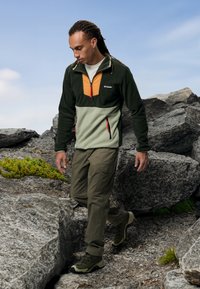 Fleece pulover u zelenoj, narančastoj i svijetlozelenoj boji s ovratnikom na patentni zatvarač; upareno s maslinastim cargo hlačama i čvrstim zelenim planinarskim čizmama.