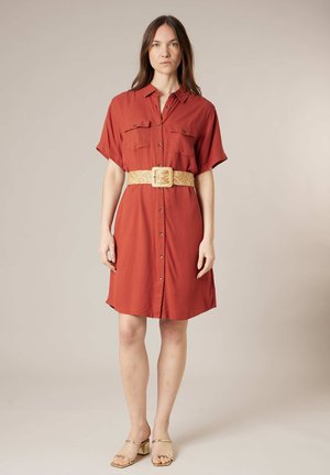 Femme debout face à l’avant portant une robe courte à manches courtes rouge rouille boutonnée avec des poches poitrine et une large ceinture beige tissée, sandales beige à talons blocs.