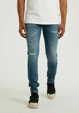 Man draagt strakzittende blauwe versleten spijkerbroek en zwarte sneakers met beige zolen, staand tegen een effen lichte achtergrond.