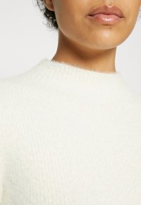 Gros plan sur le bas du visage et le cou d'une personne portant un pull blanc doux et duveteux avec un col rond.