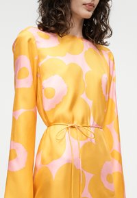 Γυναίκα που φοράει κίτρινο και ροζ σατέν φόρεμα με λουλουδάτο σχέδιο, μακριά μανίκια και λεπτή ζώνη στη μέση, με ελαφρώς καστανά σγουρά μαλλιά ορατά.