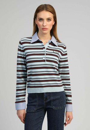 Femme aux cheveux bruns clairs et raides, portant un polo à manches longues rayé bleu, noir et rouge avec un écusson "1985", et un jean taille haute foncé.