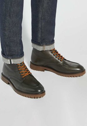 Dark green leather boots with orange laces, featuring a rugged sole and rounded toe, paired with dark jeans rolled at the cuffs.