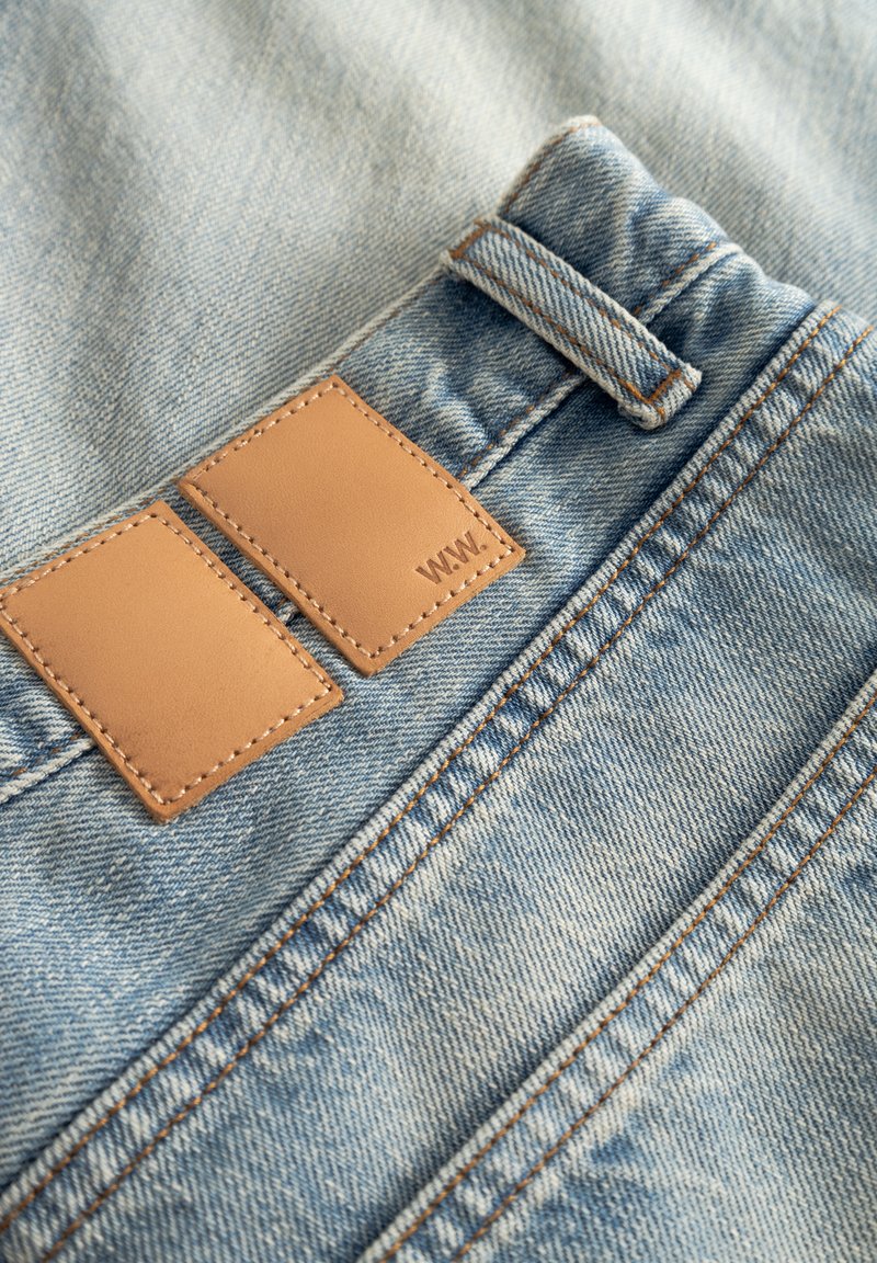 Hellblaue Denim-Stoff mit einem doppelten Stück hellbraunen Leders mit Nähten, in der Nähe eines Bunds, neben einer Gürtelschlaufe.