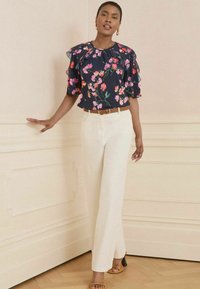 Blusa floreale blu navy con maniche a volant, abbinata a pantaloni bianchi e una cintura marrone. Il modello indossa sandali con tacco beige, in piedi davanti a una parete chiara.