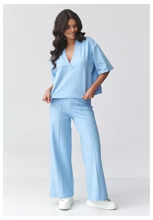 Kobieta o długich ciemnych włosach, ubrana w jasnoniebieskie szerokie spodnie, dopasowaną luźną bluzkę z dekoltem w kształcie litery V oraz białe sneakersy na platformie, uśmiechająca się i poprawiająca bluzkę.