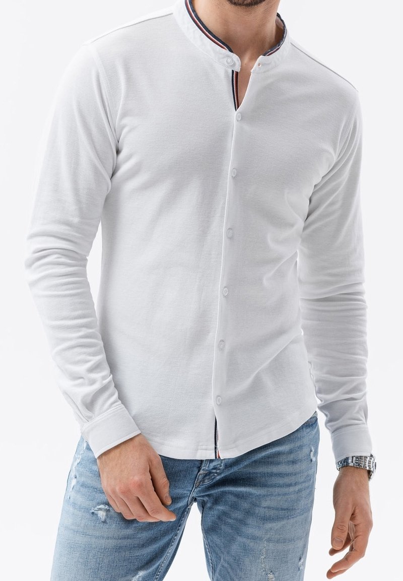 Homme portant une chemise blanche à manches longues boutonnée avec un détail de col rayé noir, blanc et rouge et un jean bleu clair déchiré.