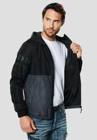 Must ja hall kapuutsiga jope lukuga, millel on tekstureeritud varrukad ja elastsed mansetid, kantud valge t-särgi peale, mis näitab elegantset disaini.