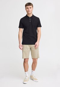 Sort polo-shirt med tre knapper, tætsiddende design, lille logo på brystet; parret med beige shorts og lyse sneakers med hvide sokker.