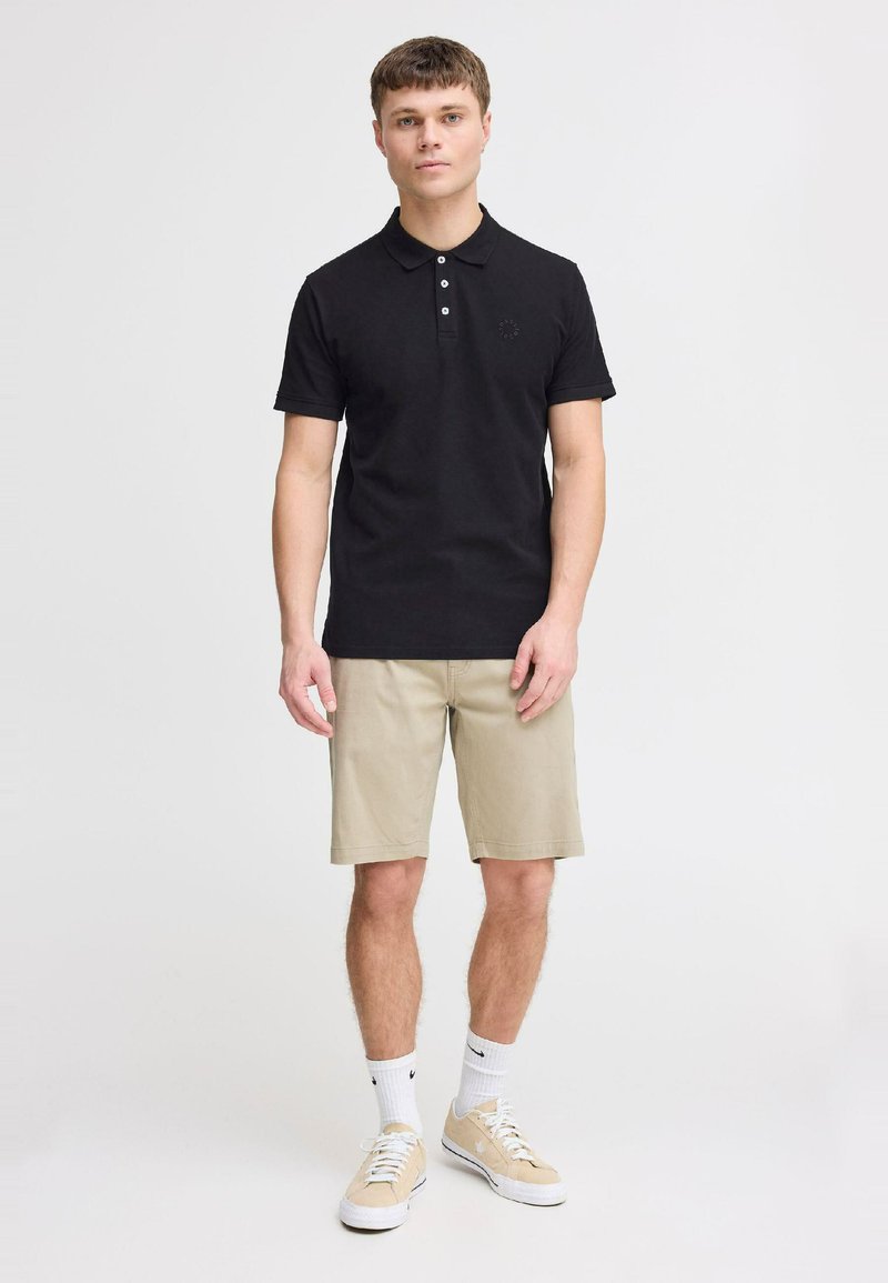 Sort polo-shirt med tre knapper, tætsiddende design, lille logo på brystet; parret med beige shorts og lyse sneakers med hvide sokker.