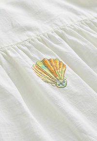 Biela látka s vyšívaným motívom mušle v oranžových, žltých a modrých niťach, so záhybom pri šve.