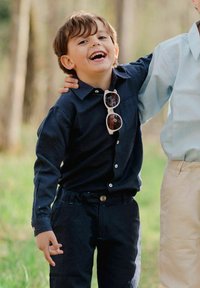 Lapsi, jolla on päällään tummansininen napitettava paita, tummat housut ja valkoiset aurinkolasit, hymyilee ja seisoo toisen henkilön kanssa käsivarsi hänen ympärillään.