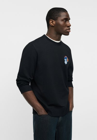 Sweat-shirt noir à col rond et poignets côtelés, arborant un logo singe coloré brodé sur la poitrine. Porté avec un jean bleu.
