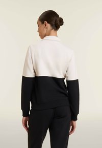 Donna con capelli scuri raccolti in uno chignon, che indossa una giacca a maniche lunghe color block in crema e nero, vista da dietro su uno sfondo semplice.