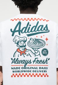 Szakács egy olyan tányért tart, amin egy Adidas tornacipő grafikája látható, a fehér ing hátulján pedig az "Always Fresh", a "Made Original Daily" és a "Worldwide Delivery" feliratok olvashatók.