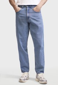 Lyseblå denimjeans med afslappet pasform, fem-lomme design og synlige syninger. Kombineret med grå og hvide sneakers.