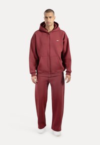 Vinrød hættetrøje med lynlås, foret hætte og forlommer, kombineret med matchende løstsiddende joggingbukser. Hvide sneakers og diskrete logodetaljer.