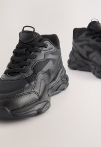 Paire de baskets noires épaisses avec des semelles épaisses et des détails texturés, présentée sur un fond clair uni.