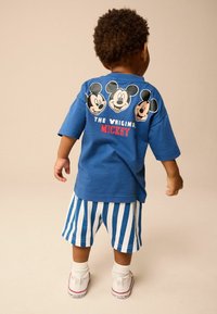 Blå t-shirt med tre Mickey Mouse-illustrationer på ryggen, parret med hvide og blå stribede shorts, iført hvide sneakers.