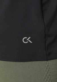 Mustaa urheilupaitaa, jossa on teksturoitu kangas, jossa on harmaa "CK"-logo alareunassa, yhdistettynä oliivinvihreisiin teksturoituihin shortsien.