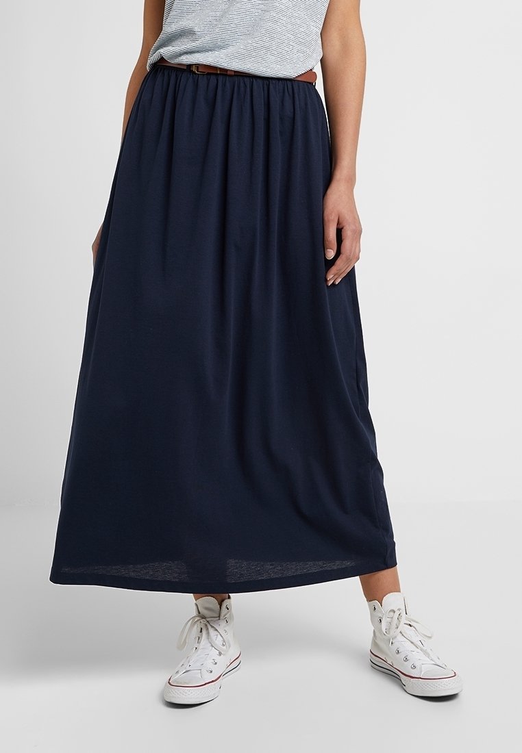 Femme portant une jupe bleu marine longueur cheville avec une ceinture marron, des baskets montantes blanches et un haut rayé gris et blanc.