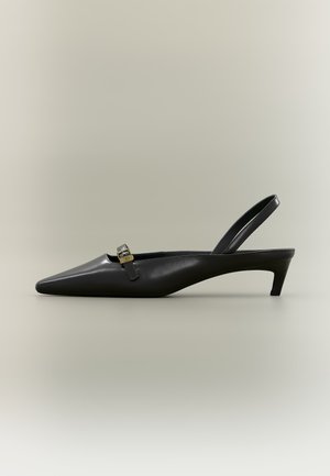 Fekete bőrből készült pántos, kis sarkú cipő hegyes orral és kis aranyszínű csattal, egyszínű háttéren.