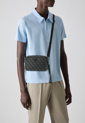 Borsa a tracolla nera con motivo, chiusura con zip, cinturino regolabile e dettaglio del logo, abbinata a una maglietta polo blu chiaro e pantaloni beige.