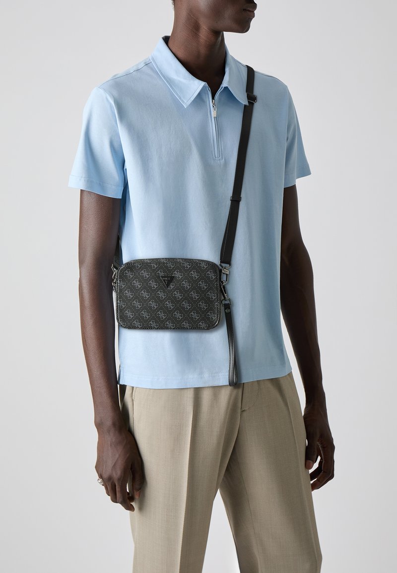 Bolso bandolero negro con estampado, cierre de cremallera, correa ajustable y detalle del logo, combinado con una camiseta polo azul claro y pantalones beige.