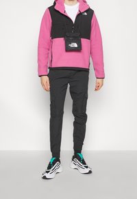 Sudadera de forro polar rosa y negra con capucha y bolsillo frontal, combinada con pantalones cargo negros y zapatillas blancas con detalles en azul verdoso.