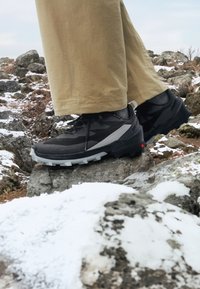 Zapatos de senderismo negros con acentos grises, suela resistente y parte superior texturizada, colocados sobre una superficie rocosa con nieve.