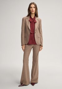 Bruin geruit pak met een op maat gemaakt blazer en uitlopende broeken. Draag een diep bordeauxrode blouse met een strik. Glanzende bordeauxrode schoenen.