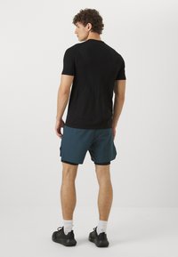 Camiseta negra de manga corta con cuello redondo, combinada con pantalones cortos deportivos en color teal. El modelo lleva zapatillas negras y calcetines blancos.