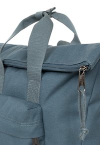 Zaino in tessuto blu con un cappuccio superiore, due manici robusti, una tasca frontale con zip e materiale testurizzato. Comprende un tirazip in metallo.
