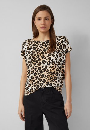 Kobieta o brązowych włosach, ubrana w krótką bluzkę w panterkę i czarne spodnie, stojąca na jednolitym jasnoszarym tle.