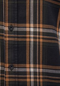 Nahaufnahme eines karierten Flanellhemdes in Schwarz, Orange und Weiß, mit einem Knopfdetail, weichem Material und strukturiertem Schnitt.