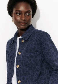 Veste en imprimé léopard bleu marine avec un col structuré, deux poches à l'avant et des boutons dorés. Tissu en coton lisse avec une texture subtile.