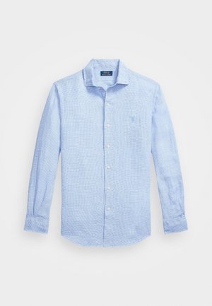 Chemise bleu clair à manches longues avec col, boutons blancs et tissu texturé subtil, avec un petit logo brodé sur la poitrine.