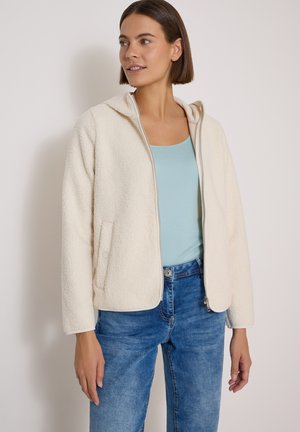 Femme portant une veste polaire crème zippée sur un débardeur bleu clair et un jean bleu sur un fond clair uni.