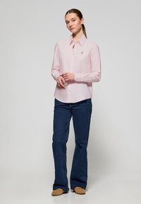 Light pink, long-sleeve button-up shirt with a collar and embroidered logo. Paired with dark blue, flared jeans and beige shoes.