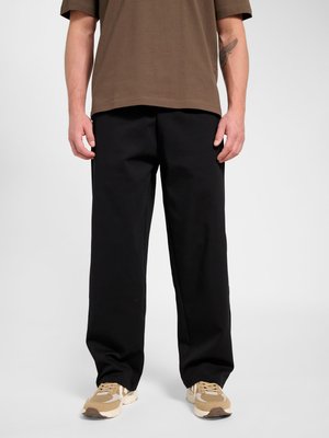 Mann trägt lockere schwarze Hose, beige und weiße Sneaker, und ein braunes kurzärmeliges Hemd steht vor einfarbigem Hintergrund.
