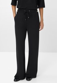 Schwarze weit geschnittene Hose aus glattem Stoff, mit einem Kordelzugbund, seitlichen Taschen und metallischen Hardware-Akzenten.